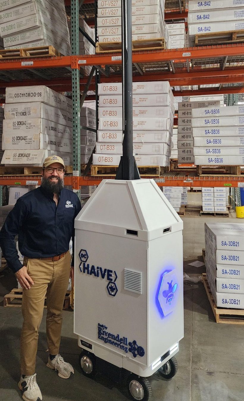Craig Olivo standing beside the DC HAiVE rover in a distribution center aisle. The machine is about chest height with its camera mast rising above it, marked DC HAiVE and Built by Rivendell Engineering.