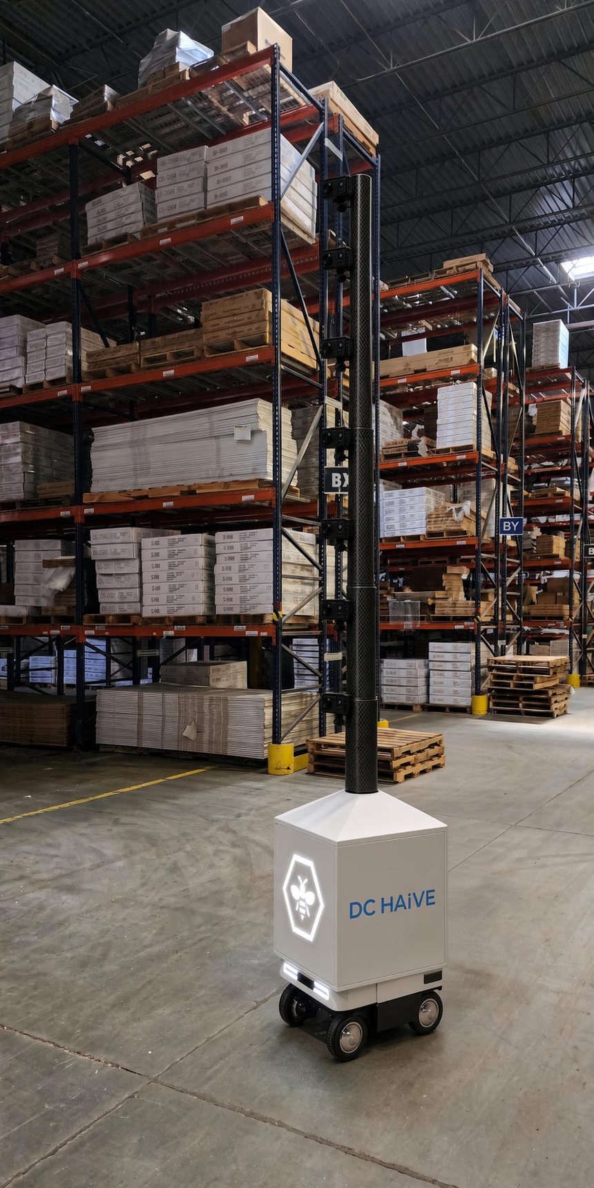 The DC HAiVE rover standing in a distribution center aisle — a compact white four-wheeled ground vehicle, roughly waist height, carrying a slim carbon-fiber mast that rises past the top rack level.